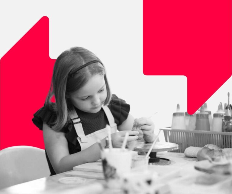 Young girl painting while sat at a table. There are paintbrushes and pots of paints on the table in front of her.